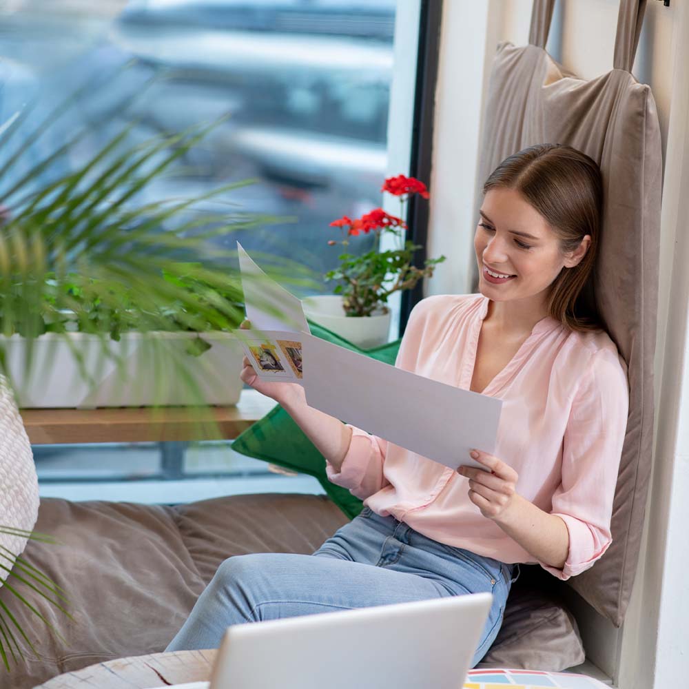 Young woman sitting by a window, reading documents with a smile, surrounded by greenery and natural light, symbolizing reflection and positivity – an embodiment of hope and calm.